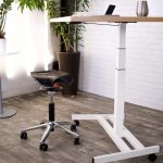 White standing desk with a wood-tone surface beside a stool and plants